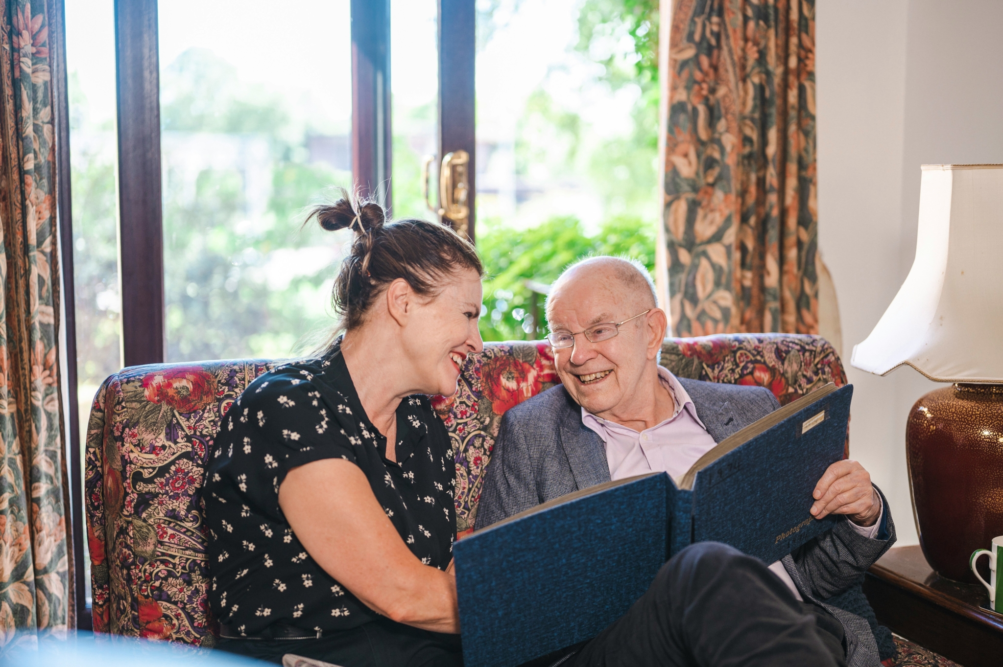 A woman and a senior gentleman sat on a sofa, smiling and looking through a large book.